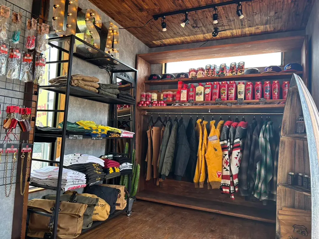 インダストリアルスタイルの店舗内装。黒いスチール棚に折りたたまれた衣類、木製フレームのクローゼットに吊るされたジャケットやシャツ、上部棚に並ぶ赤いパッケージの商品が陳列されている